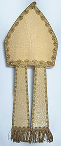 Studio photograph of the bishop's miter, a pointed cream-colored headpiece with fabric strips that extend down from the bottom edge.